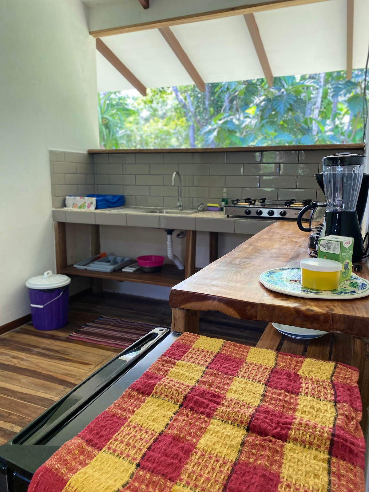 The kitchen features wooden countertops and a modern gas stove, with a blender and utensils visible. Greenery outside the window enhances the natural light that fills the space. A dish rack is present, and a bin is located beneath the counter.