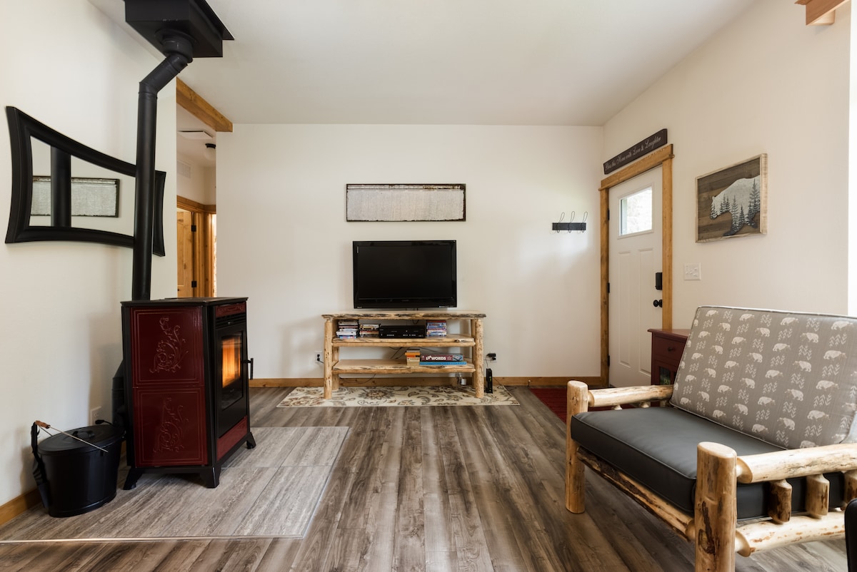 An inviting living area features a stylish wood stove and a flat-screen TV on a rustic console. A log-style sofa is positioned nearby, complementing the natural wood accents. The floor showcases wide, light-colored planks, enhancing the airy feel of the space.