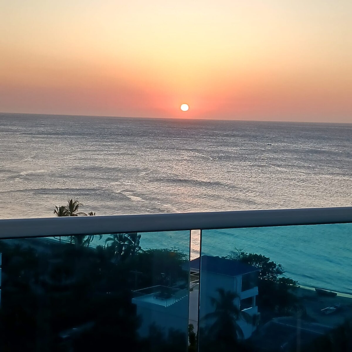 A stunning sunset is captured over the ocean, with the sun hovering just above the horizon. The sky displays warm hues of orange and pink, reflecting on the water. A glass railing offers an unobstructed view of the serene scene.
