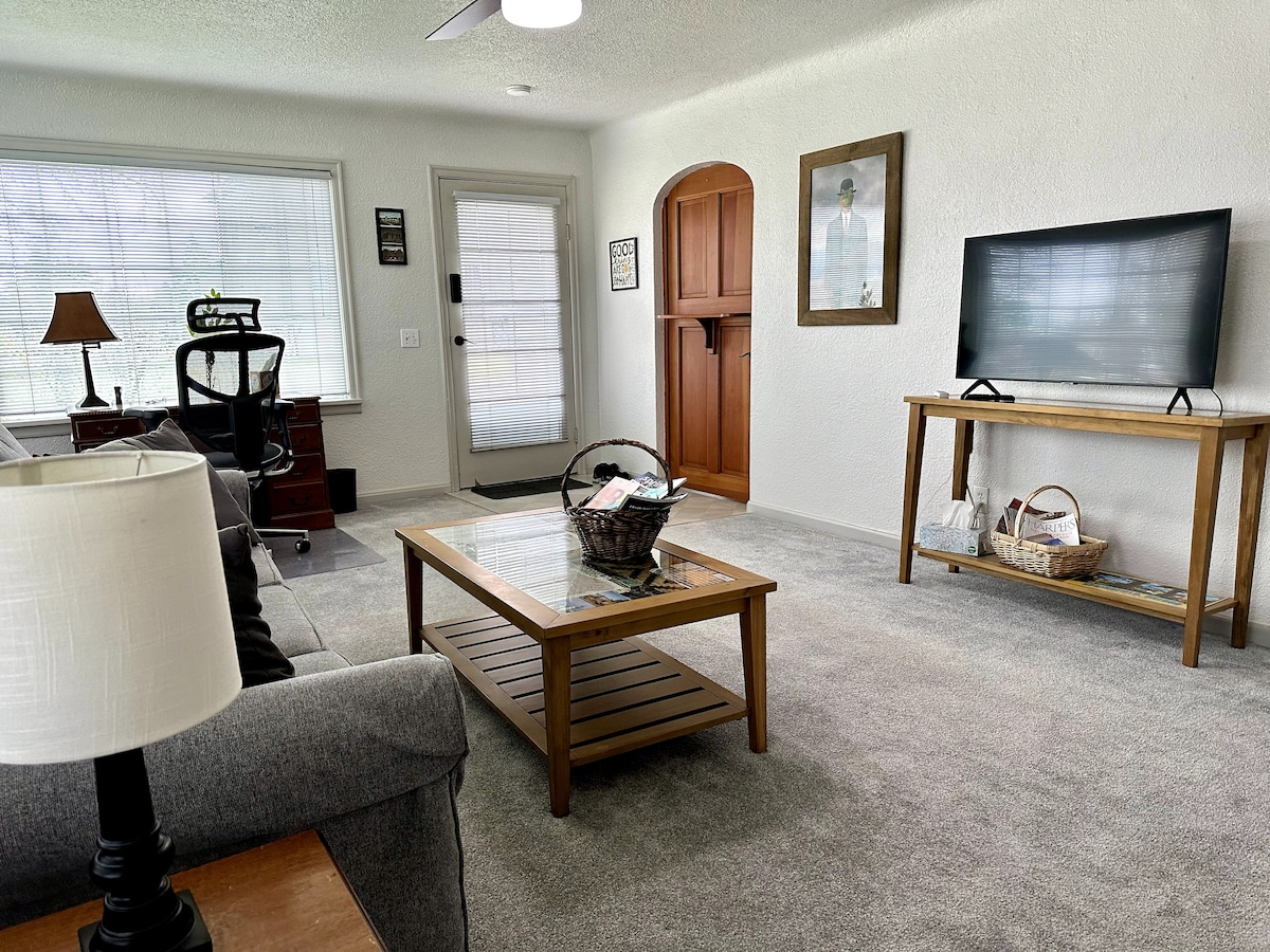 The living room features a soft carpet underfoot and natural light streaming through large windows. A comfortable seating area is arranged around a wooden coffee table, with a Smart TV on a stand. A desk and chair are positioned near the door, providing a workspace.