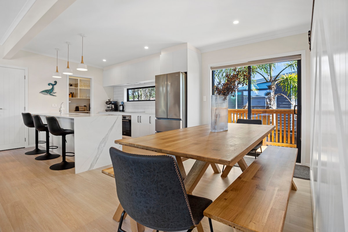 An open plan kitchen and dining area is visible, featuring a wooden dining table with attached benches. Four bar stools are arranged at the kitchen island, which includes a marble countertop. Large windows allow natural light and offer a view of the outdoor deck.
