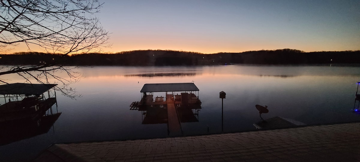 Dawn breaks over Lake of the Ozarks, with soft hues illuminating the horizon. The calm water reflects silhouettes of nearby docks, while a peaceful stillness envelops the scene. An inviting dock leads into the lake, enhancing the serene atmosphere of the surrounding landscape.