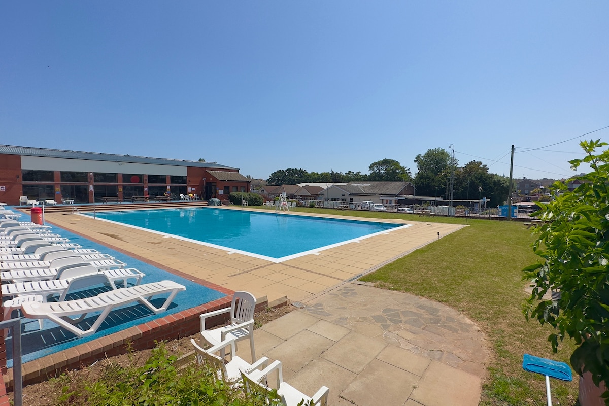 An outdoor swimming pool is surrounded by a spacious tiled deck, featuring numerous lounge chairs. Lush green grass is visible along the edges, with clear blue skies overhead. The pool area is well-maintained, inviting relaxation and enjoyment during sunny days.