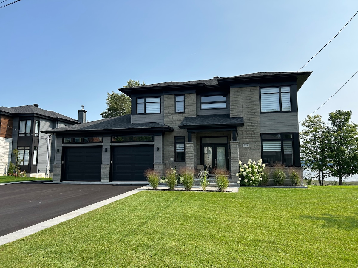 A modern two-story home features a clean façade with a combination of stone and siding. Large windows allow for natural light, while a landscaped yard offers green grass and well-maintained shrubs. A paved driveway leads up to a double garage, enhancing the home's curb appeal.