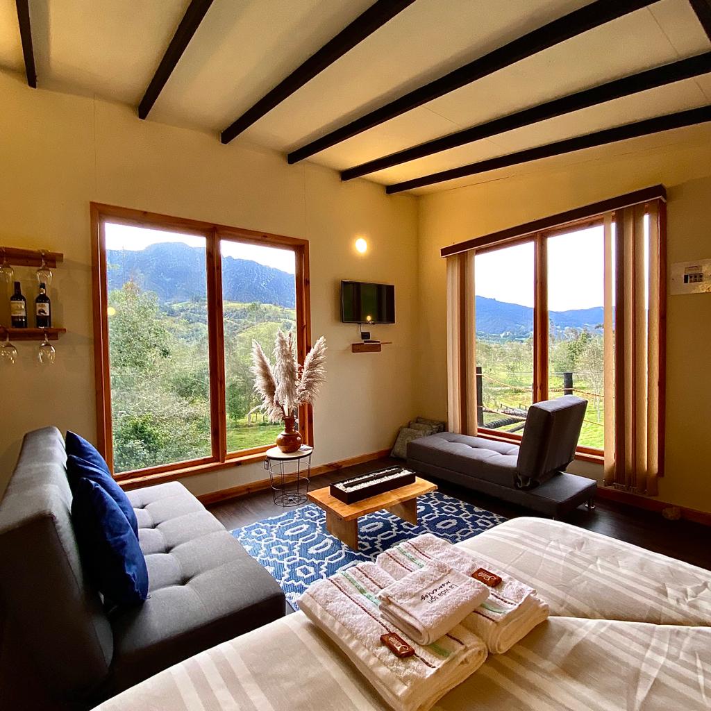 The interior features two sofas positioned near large windows that offer expansive views of the surrounding mountains. A small wooden table sits in the center, and decorative plants enhance the serene environment. The space is bright and welcoming with natural light flowing in.