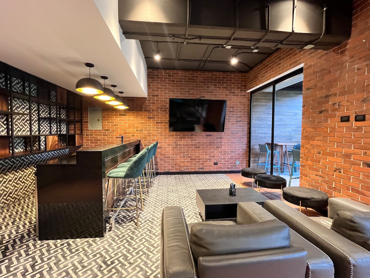 A modern lounge area is featured with exposed brick walls and stylish black furnishings. Bar stools with green upholstery surround a long counter. A large television is mounted on the wall, and large glass doors provide access to an outdoor space.