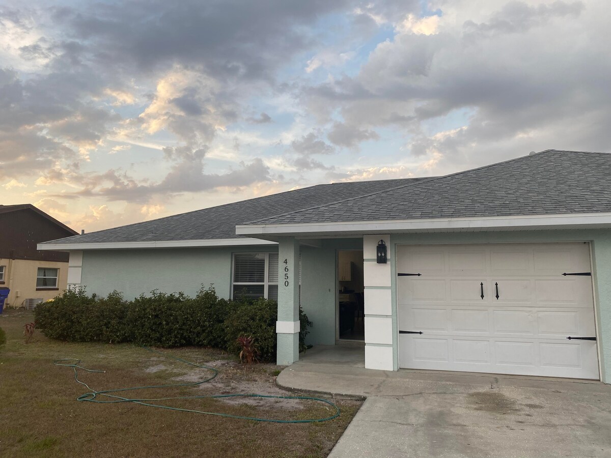 The exterior of the villa is captured during golden hour, featuring a light blue facade and a well-maintained lawn. A driveway leads to a spacious garage with contrasting black hardware. A partly cloudy sky enhances the serene ambiance surrounding the property.