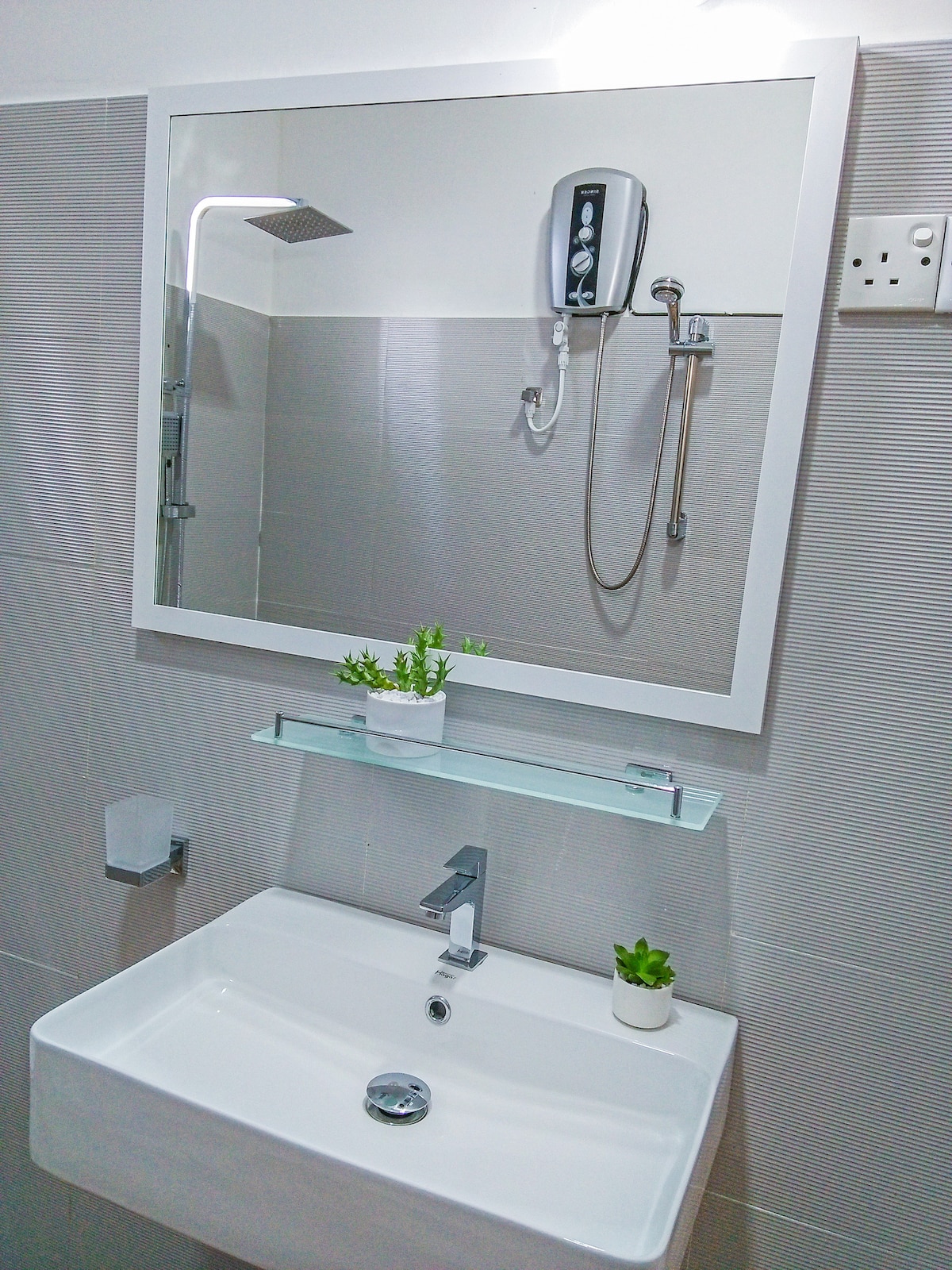 A modern bathroom features a sleek, white sink with a minimalist design. Above the sink, a large mirror reflects the space, while a shower is visible with a handheld faucet. Small potted plants add a touch of greenery on a glass shelf.