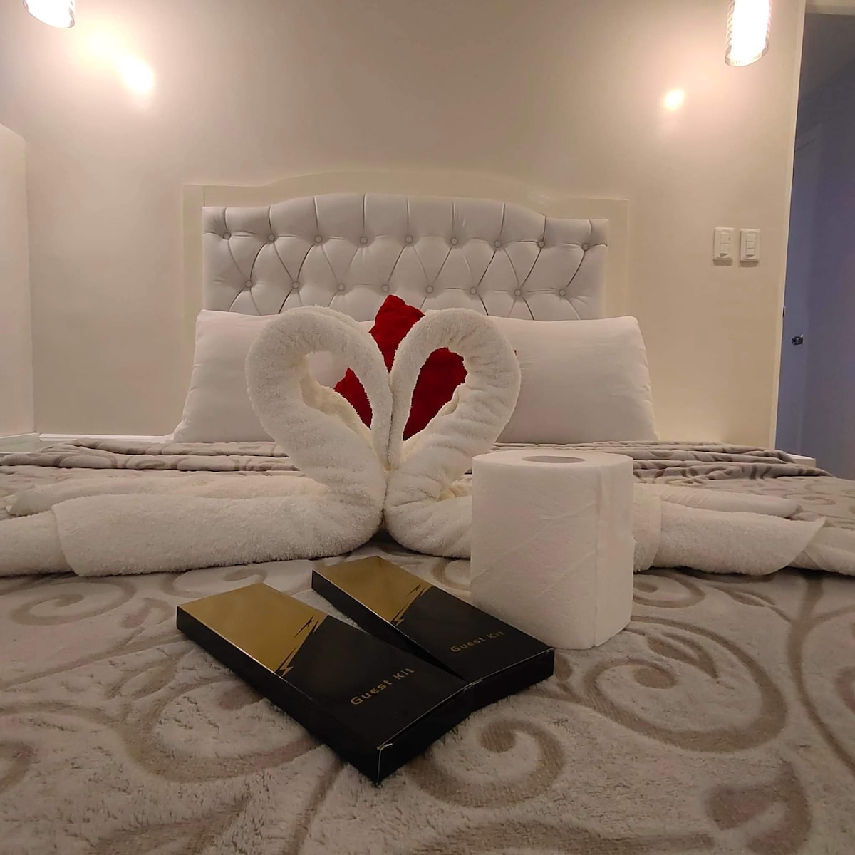 A neatly arranged bed displays a white headboard against a light-colored wall. Two white towels are folded into a heart shape on the bed, complemented by decorative red accents. A roll of toilet paper and a box of tissues are placed on a soft, patterned blanket.