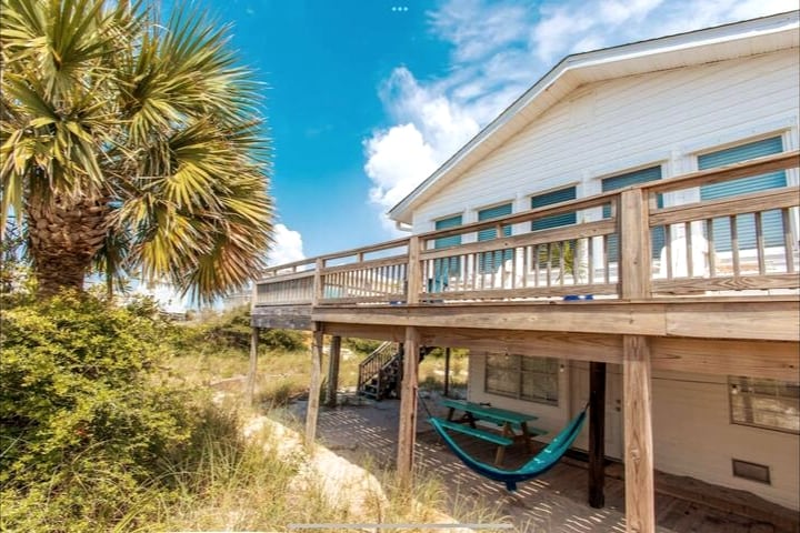 A spacious outdoor deck is visible, featuring a colorful hammock suspended between posts. Lush, green vegetation surrounds the area, with palm trees framing the scene and blue skies above.