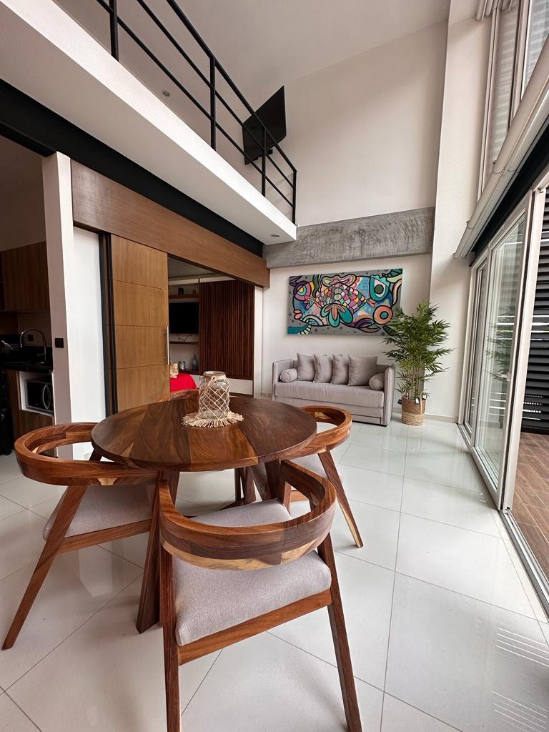 An open dining area features a round wooden table surrounded by four curved chairs with soft cushions. A colorful piece of wall art is displayed above a small grey sofa, while large glass doors lead to an outdoor space, allowing natural light to fill the room.