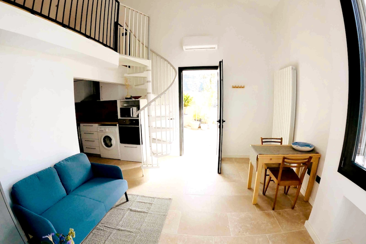A bright and airy living area features a blue sofa and a wooden dining table with chairs, positioned beside a large door leading to an outdoor space. A spiral staircase is visible in the background, with an open kitchen area seen to the left.