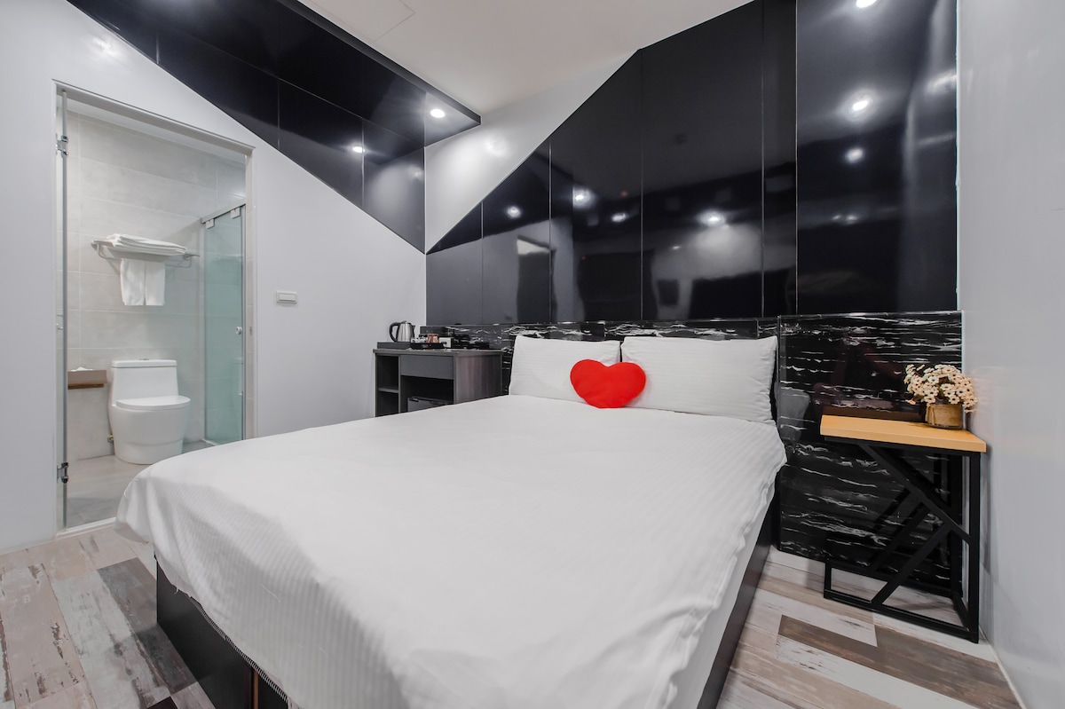 A modern sleeping area features a spacious bed centered in the room, accented by a red heart-shaped decor piece. A small table with decorative flowers is included. A bathroom area is visible through an open door, showcasing minimalist design elements.