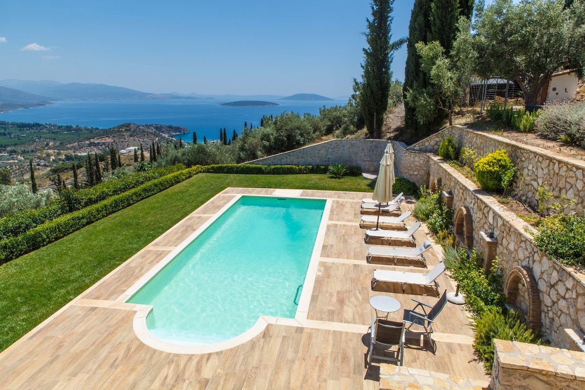A large outdoor swimming pool is set against a backdrop of lush gardens and panoramic views of the sea and mountains. Sun loungers are arranged around the pool area, providing a spacious and inviting place to relax in a tranquil environment.