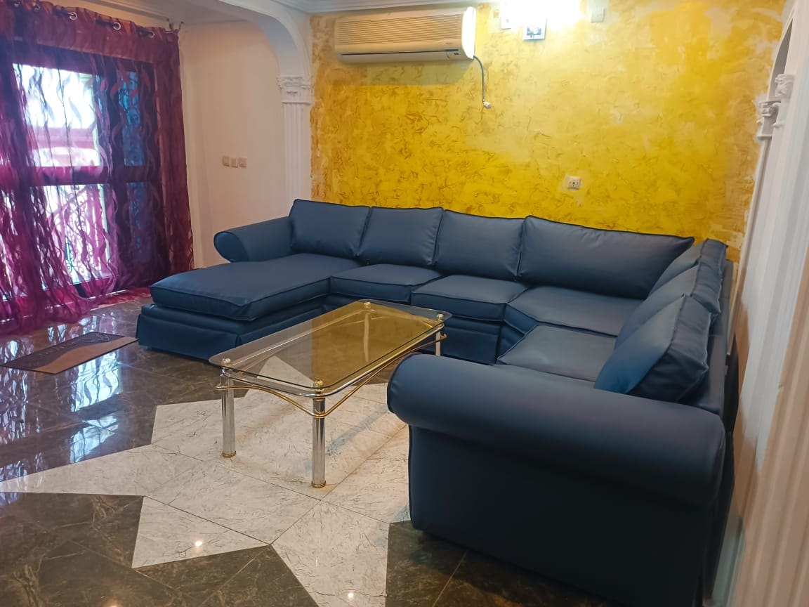 A spacious living area features a large blue sectional sofa, providing ample seating. A glass coffee table is centered on a polished marble floor. Natural light filters through windows dressed with sheer curtains, and an air conditioning unit is mounted on the wall.