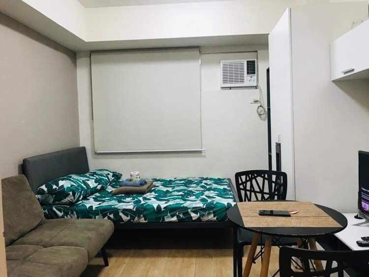A cozy room is furnished with a spacious bed, adorned with leafy patterned bedding, and a sofa bed nearby. A round table with a woven placemat sits beside a wall-mounted television. Natural light filters through a shaded window, enhancing the comfortable environment.