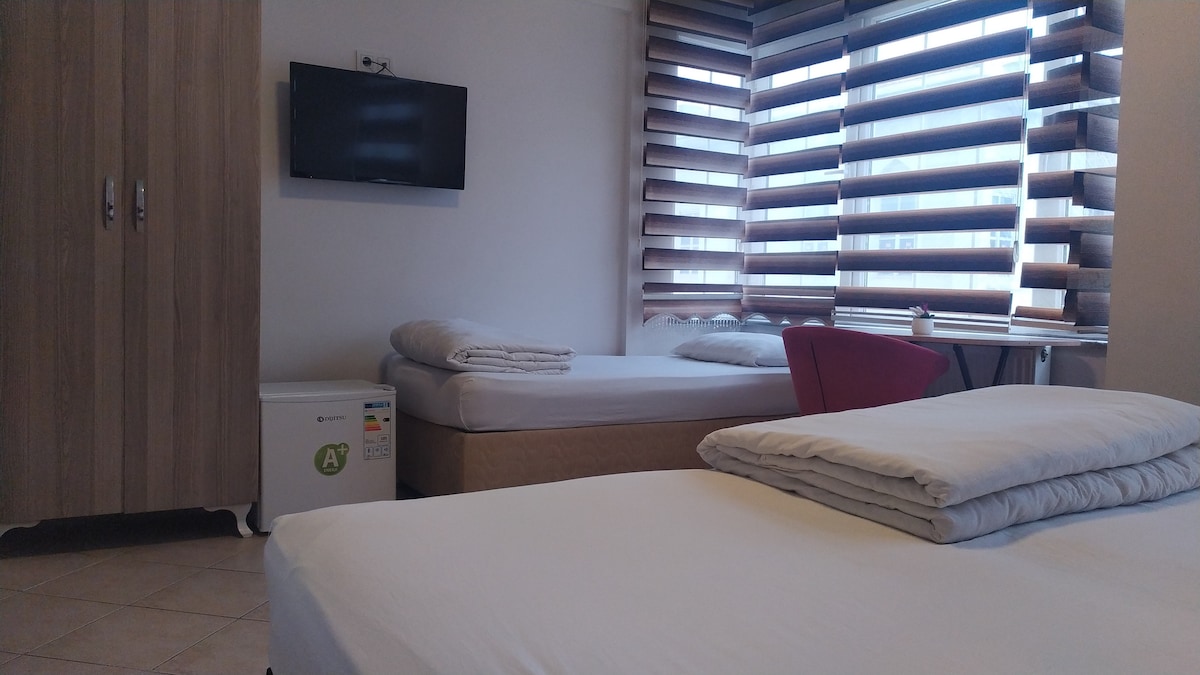 A well-lit room features two spacious beds with neutral bedding. A wall-mounted television is present, alongside a wooden wardrobe. Large windows with horizontal blinds allow natural light to filter in, while a small refrigerator is visible in the corner.