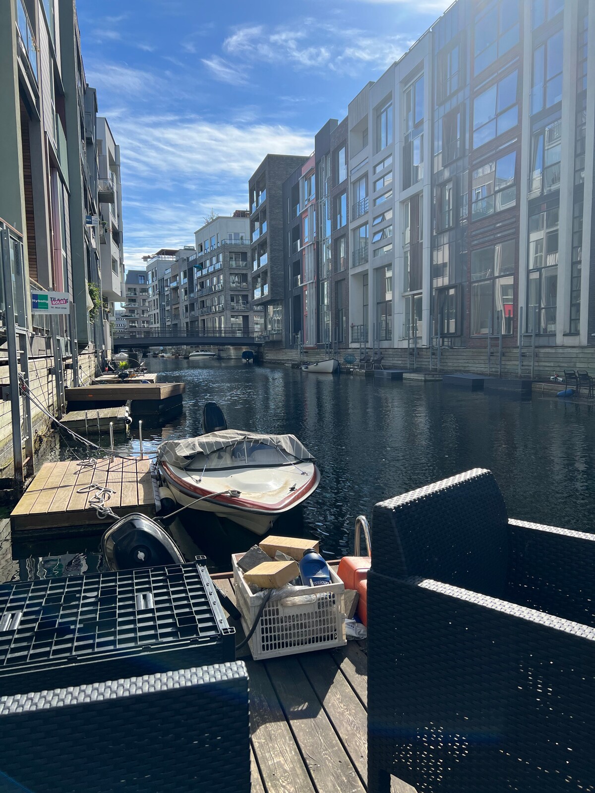 A serene canal view showcases modern buildings lining the waterway. A small boat is moored alongside a wooden dock, which features seating for relaxation. Sunlight reflects off the water, enhancing the tranquil atmosphere of the area.