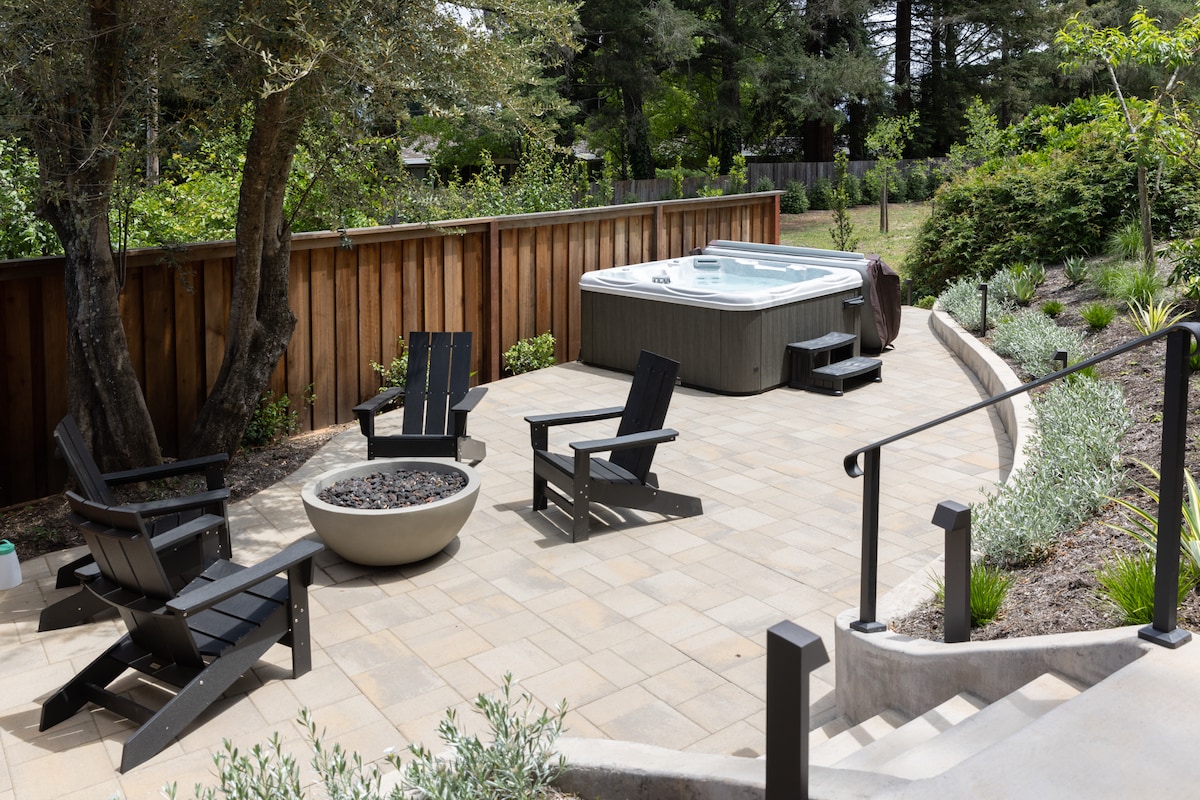 An outdoor space features a hot tub surrounded by four black lounge chairs and a circular fire pit. The area is paved with large stone tiles and bordered by greenery and wooden fencing, creating a private and relaxing atmosphere.