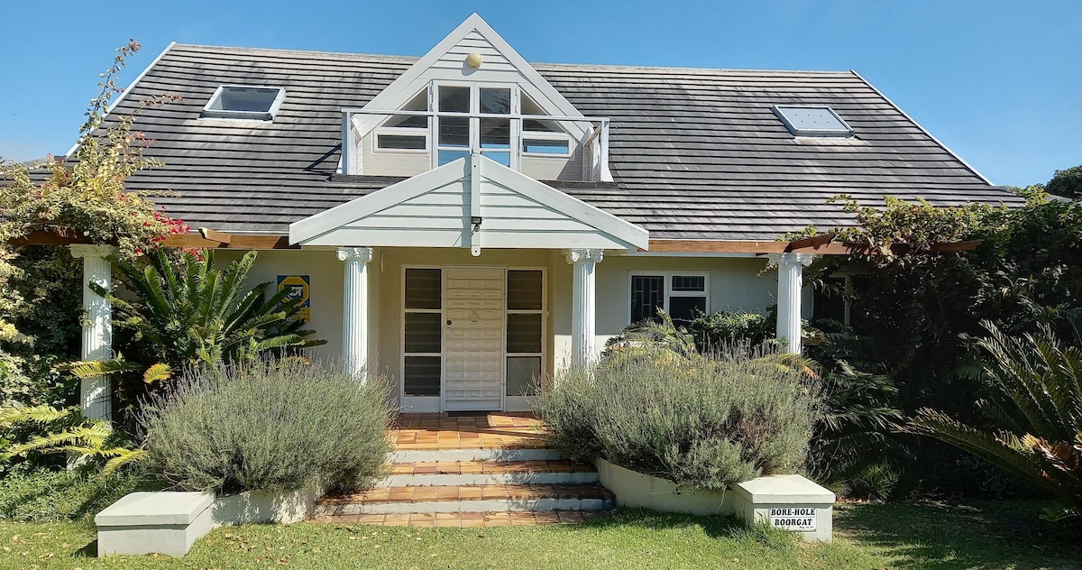The villa's exterior is presented, featuring a symmetrical design with a sloped roof and large front windows. Lush greenery and lavender plants frame the entrance, which leads to a set of double doors. The pathway is bordered by a manicured lawn and small stone steps.