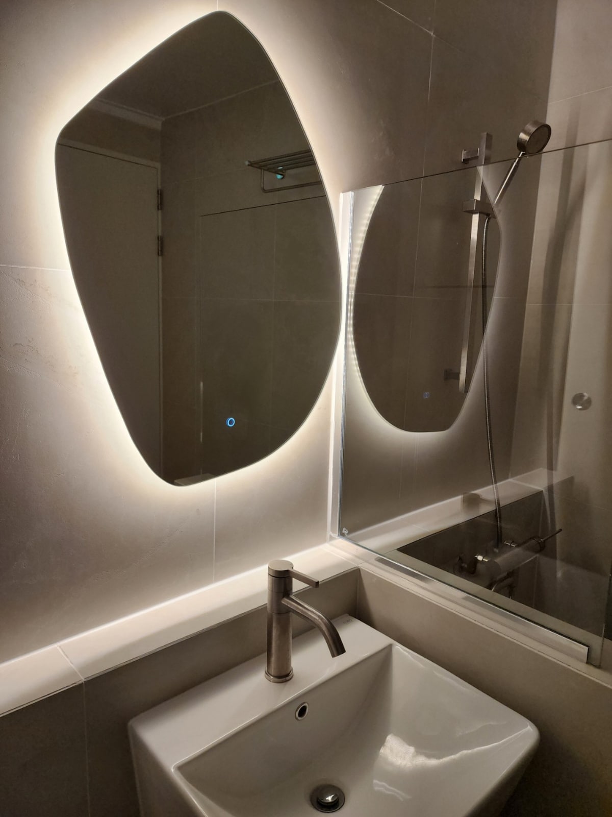 A modern bathroom features a uniquely shaped mirror illuminated by soft lighting. A sleek sink is positioned below the mirror, complemented by a contemporary faucet. Light-colored tiles create a clean and spacious appearance, while a showerhead is visible in the background.
