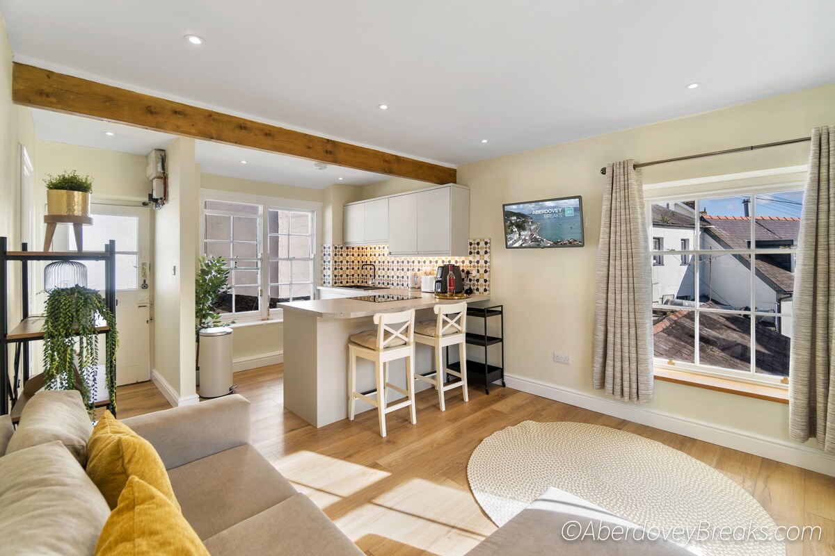 An open-plan kitchen and lounge area is depicted, featuring a central island with bar stools and modern appliances. Natural light floods through the windows, illuminating the neutral-toned walls and warm wooden accents. A comfortable sofa is positioned nearby, enhancing the cozy ambiance.