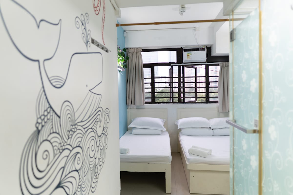 Two comfortable beds are arranged in a light-blue room, featuring a playful whale mural on one wall. Natural light enters through a window with horizontal slats, and air conditioning is visible above. Fresh linens are neatly placed on each bed.