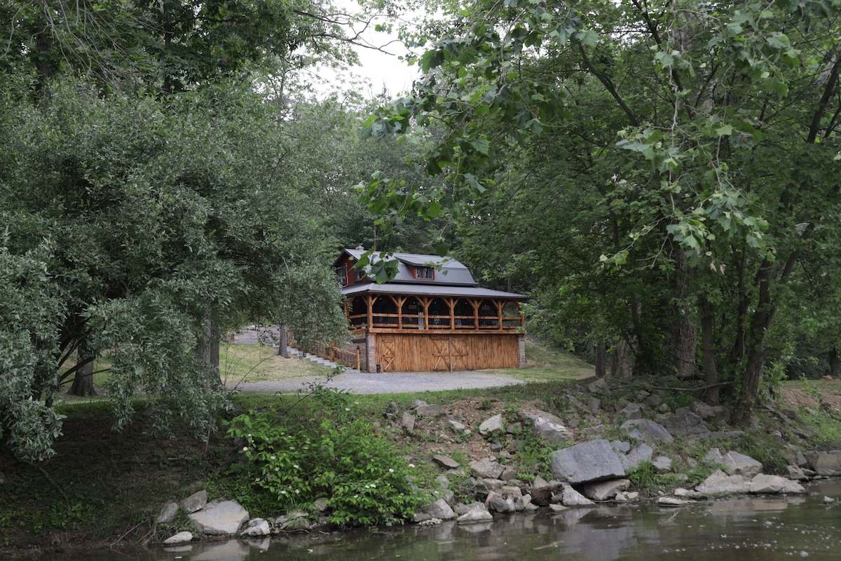 A rustic cabin is nestled among lush trees, overlooking a serene creek. The spacious porch is visible, providing outdoor seating areas with a natural backdrop. Smooth stones line the creek's edge, enhancing the tranquil setting.