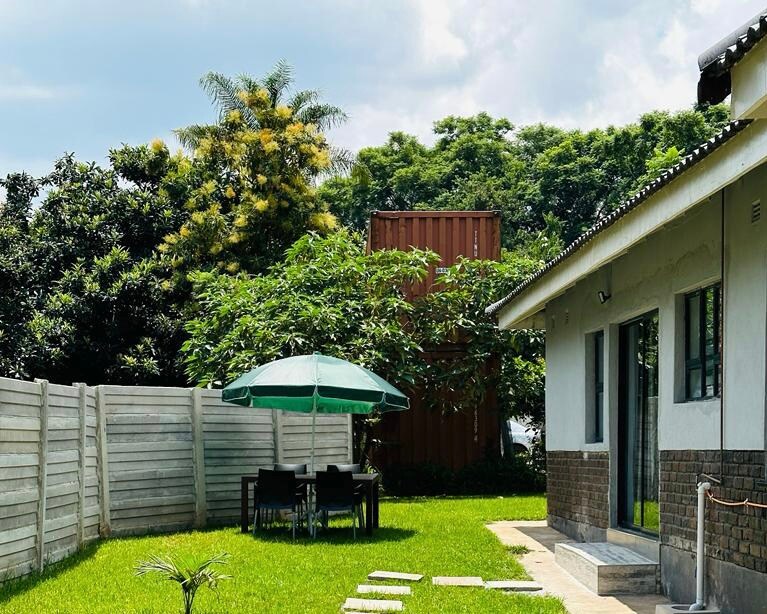 A spacious outdoor area features a green lawn under a bright sky. A round table with four chairs is sheltered by a green umbrella, surrounded by trees and a wooden fence. The structure of the studio is visible to one side.