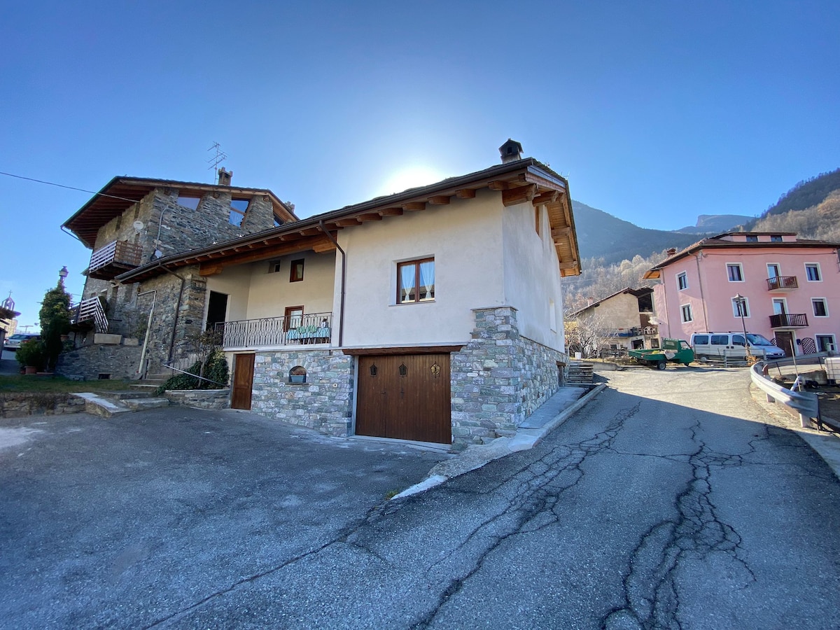 A charming building is shown, featuring natural stone and wood accents. The entrance is marked by a wooden door, and the surrounding area is paved with asphalt. Mountains form a scenic backdrop, adding to the tranquility of the location.