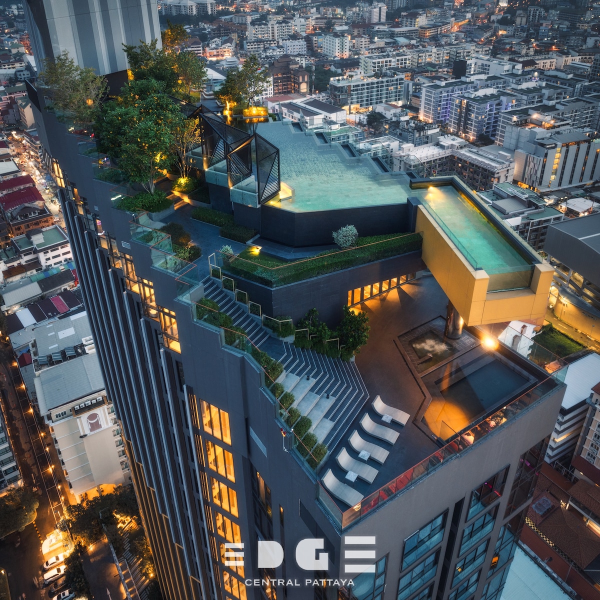 An aerial view captures the rooftop pool and terrace area of EDGE Central Pattaya. Lounge chairs are arranged near the edge, and greenery adds a touch of nature. The city skyline illuminates the background, showcasing the vibrant atmosphere of Pattaya.