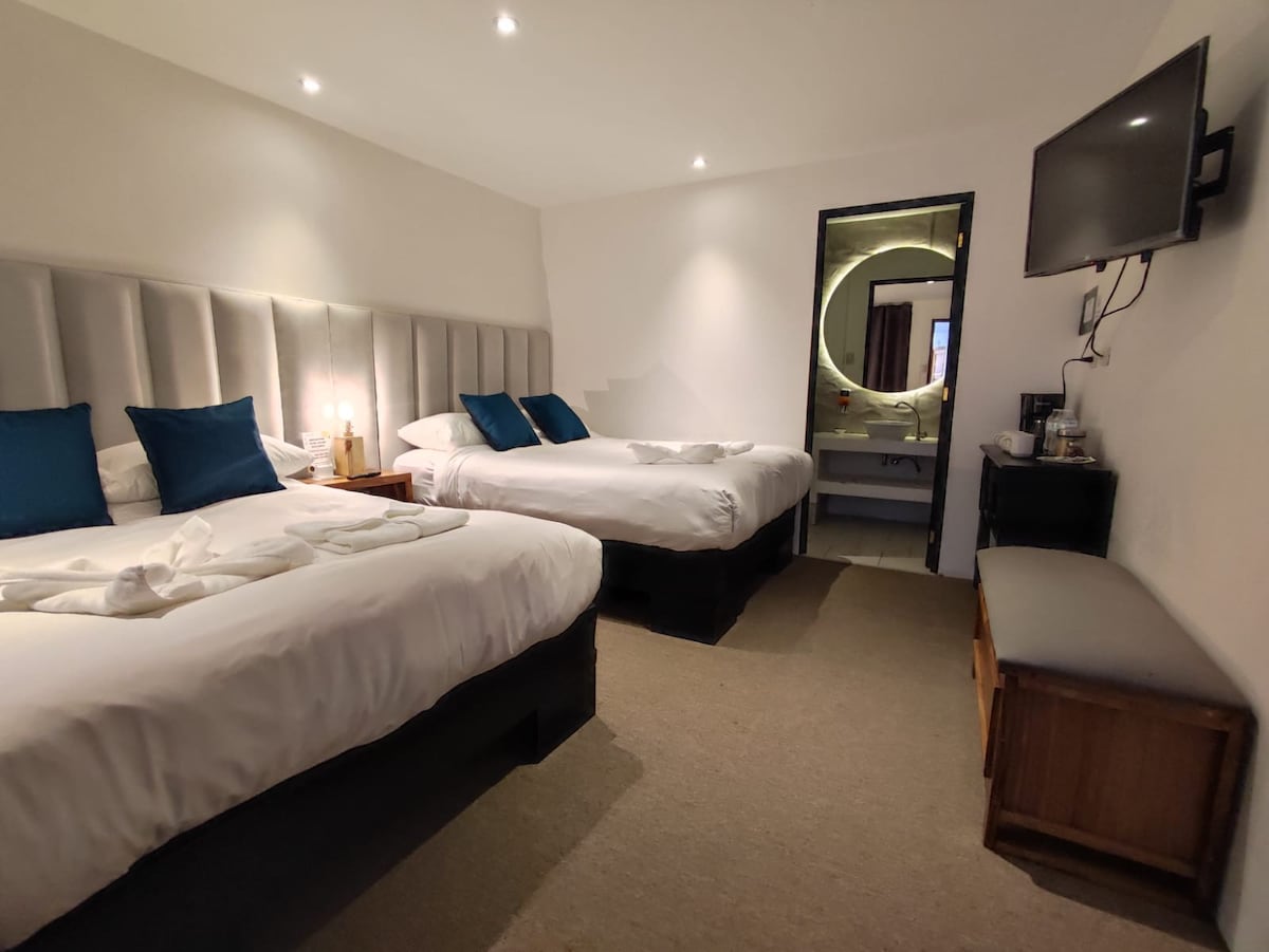 A spacious room is furnished with two comfortable beds dressed in white linens and accented by blue throw pillows. Soft lighting is provided by wall-mounted lamps. A TV is mounted on the wall, and a seating area is available for relaxation. A bathroom is visible in the background.