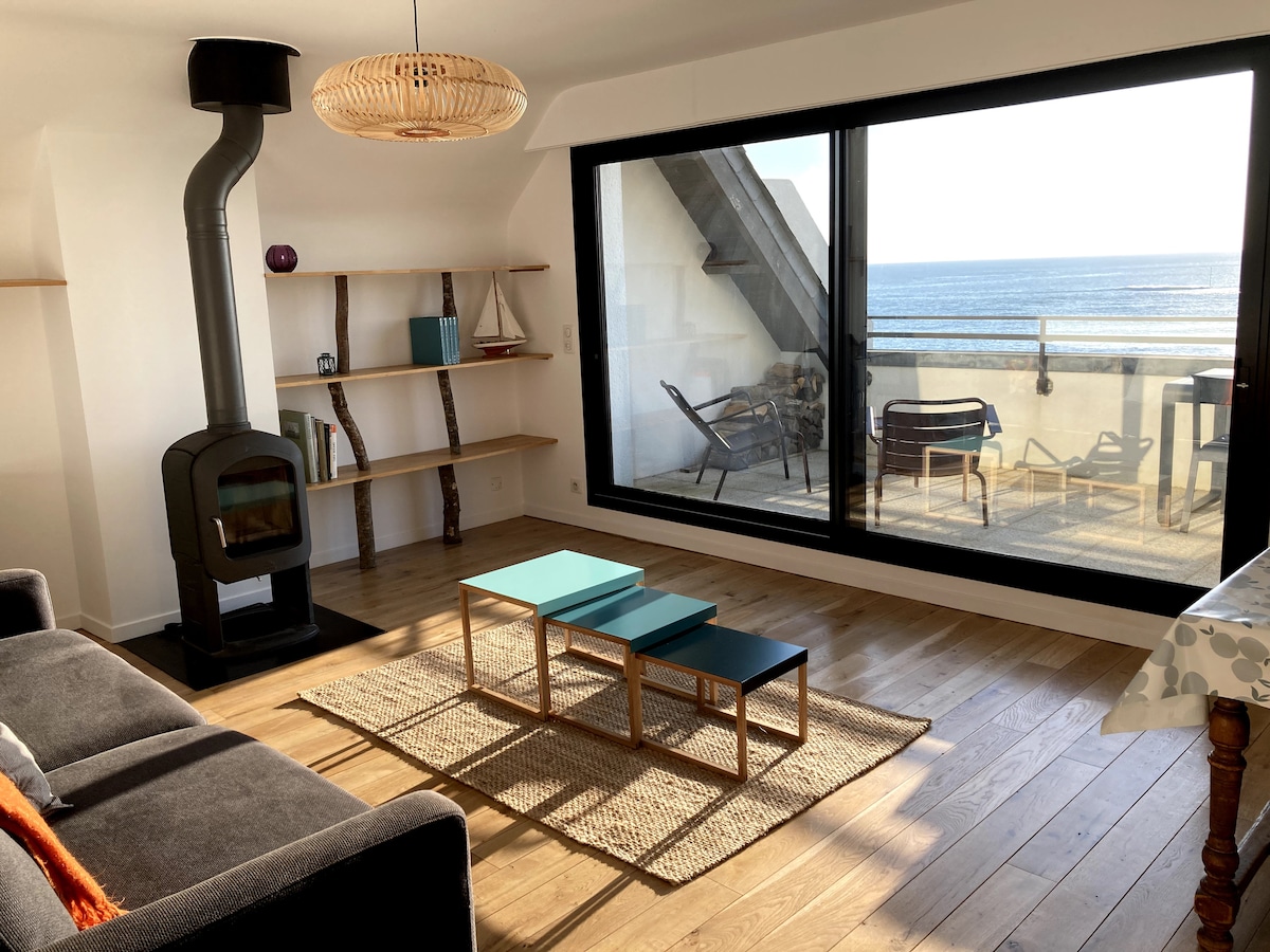 A welcoming living area showcases a comfortable sofa and a modern coffee table set. A large glass sliding door offers views of the ocean and access to a terrace. A wood stove is positioned beside a shelving unit, enhancing the space's functionality.