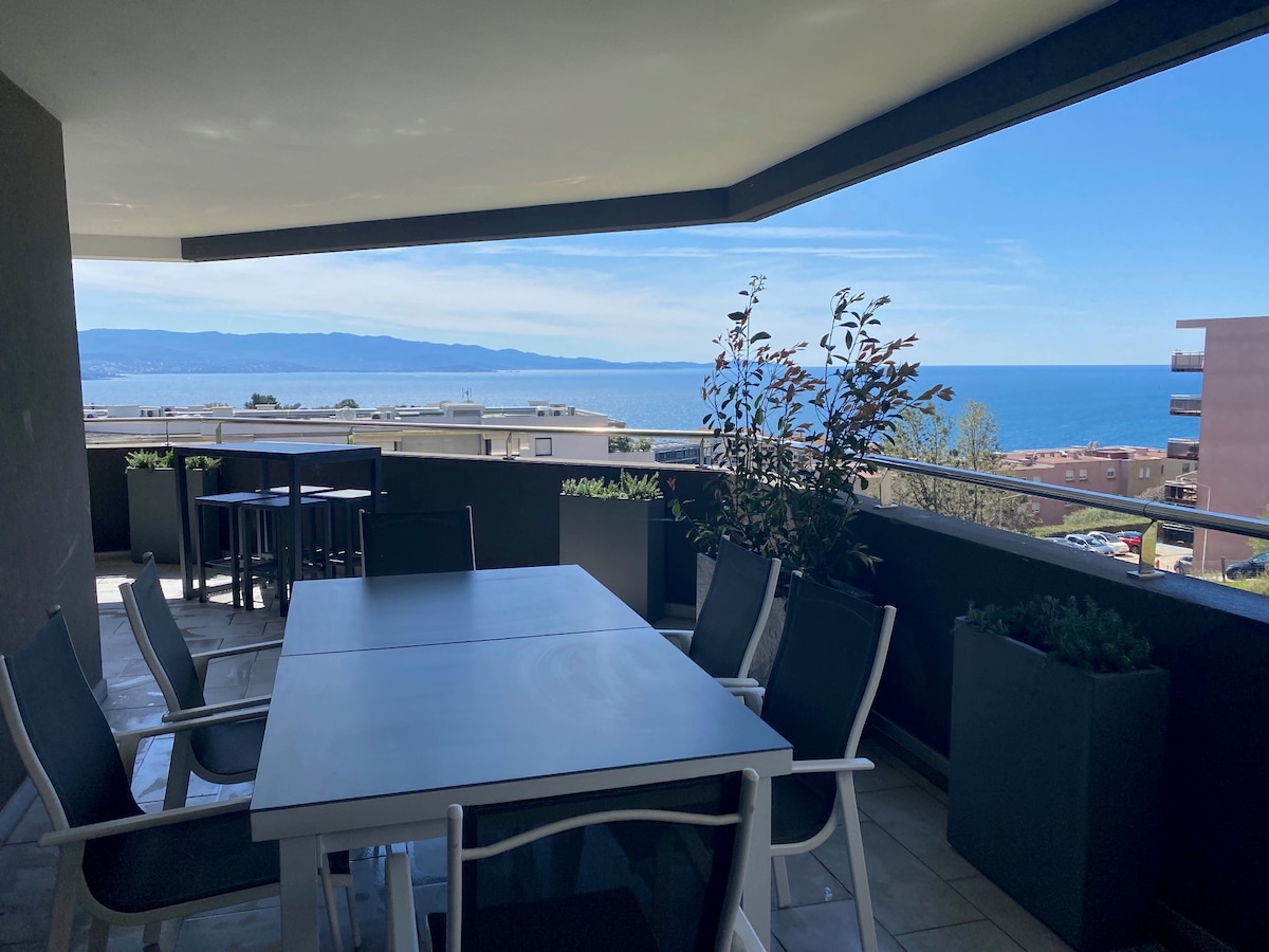 A spacious terrace is seen, furnished with a large table and chairs, providing a panoramic view of the sea and distant mountains. Planters with greenery add a natural touch to the setting, which is designed for relaxation and outdoor dining.