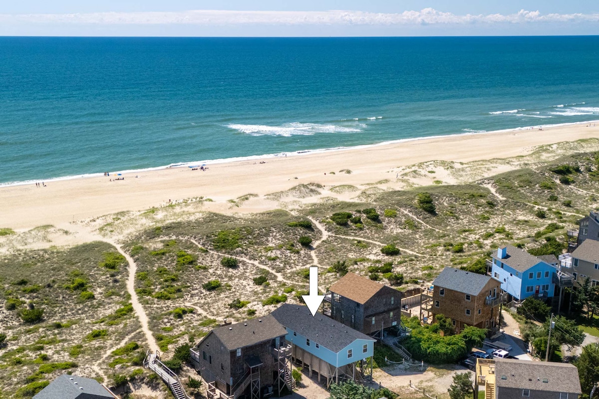 An aerial view highlights a beachside property surrounded by open sand dunes and a blue ocean. The expansive beach stretches into the distance, inviting outdoor activities. The house is framed by other residences, emphasizing its proximity to coastal amenities and the ocean.
