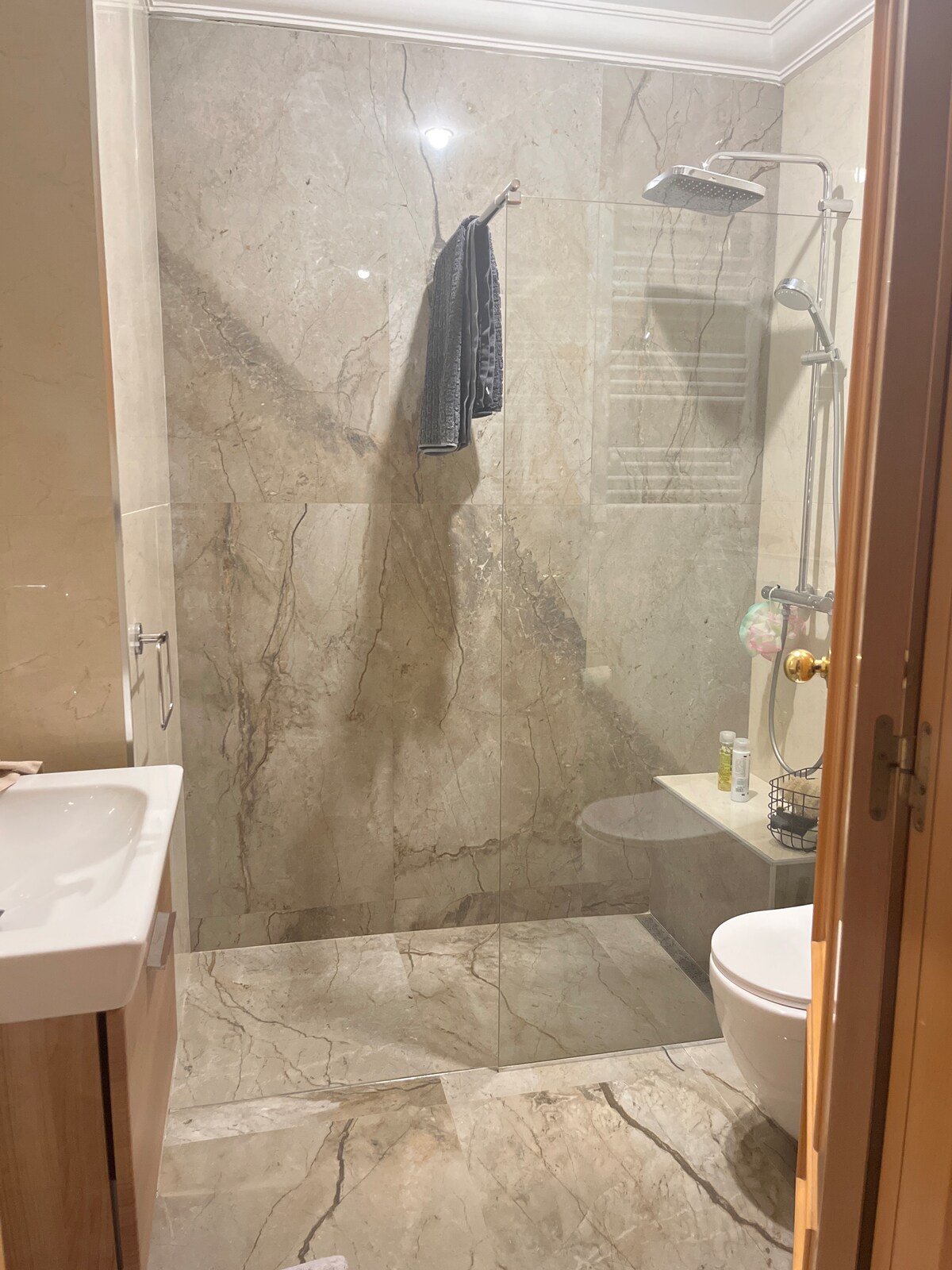 A modern bathroom features a glass shower enclosure with elegant stone wall tiles. A wall-mounted showerhead is visible, along with a sleek sink and toilet. Natural light highlights the spacious layout, while a towel hangs neatly on the glass. Subtle decor accents are present.