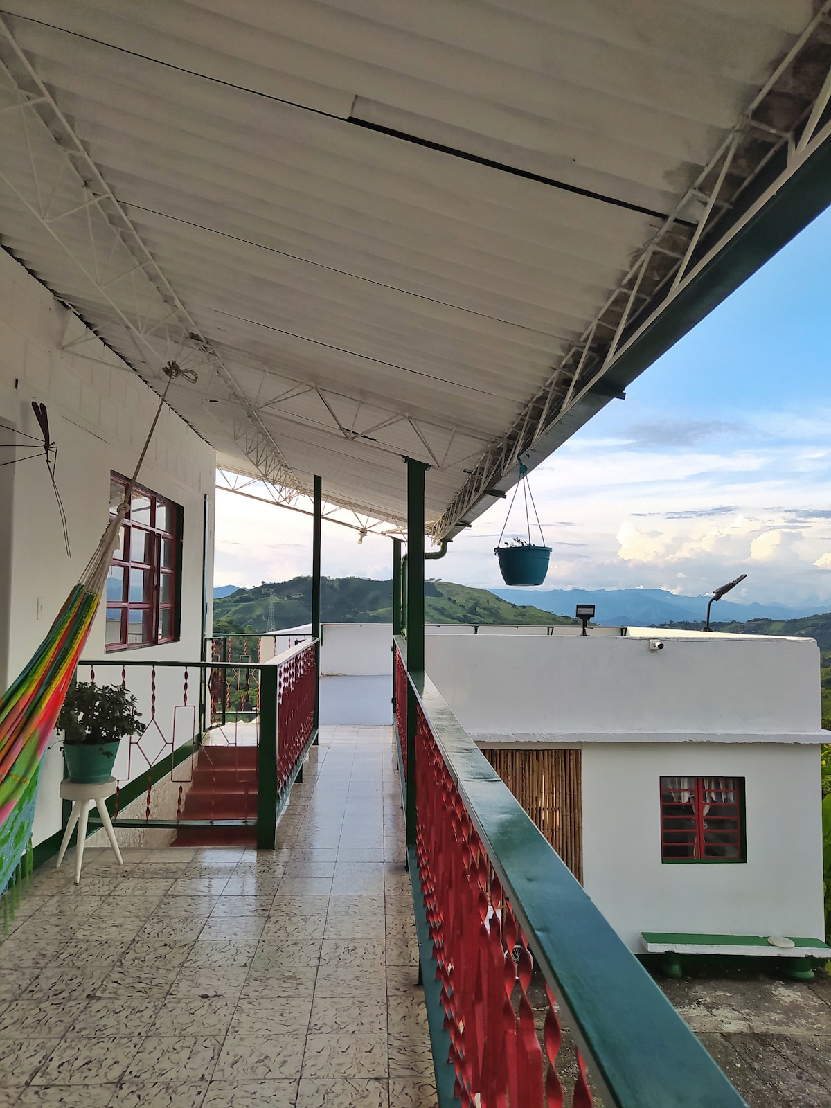 A covered balcony extends along the building, offering a panoramic view of the mountains. Colorful hammocks are draped alongside the railing, and potted plants add a touch of greenery. The floor features a patterned tile design, contributing to the warm, inviting ambiance.