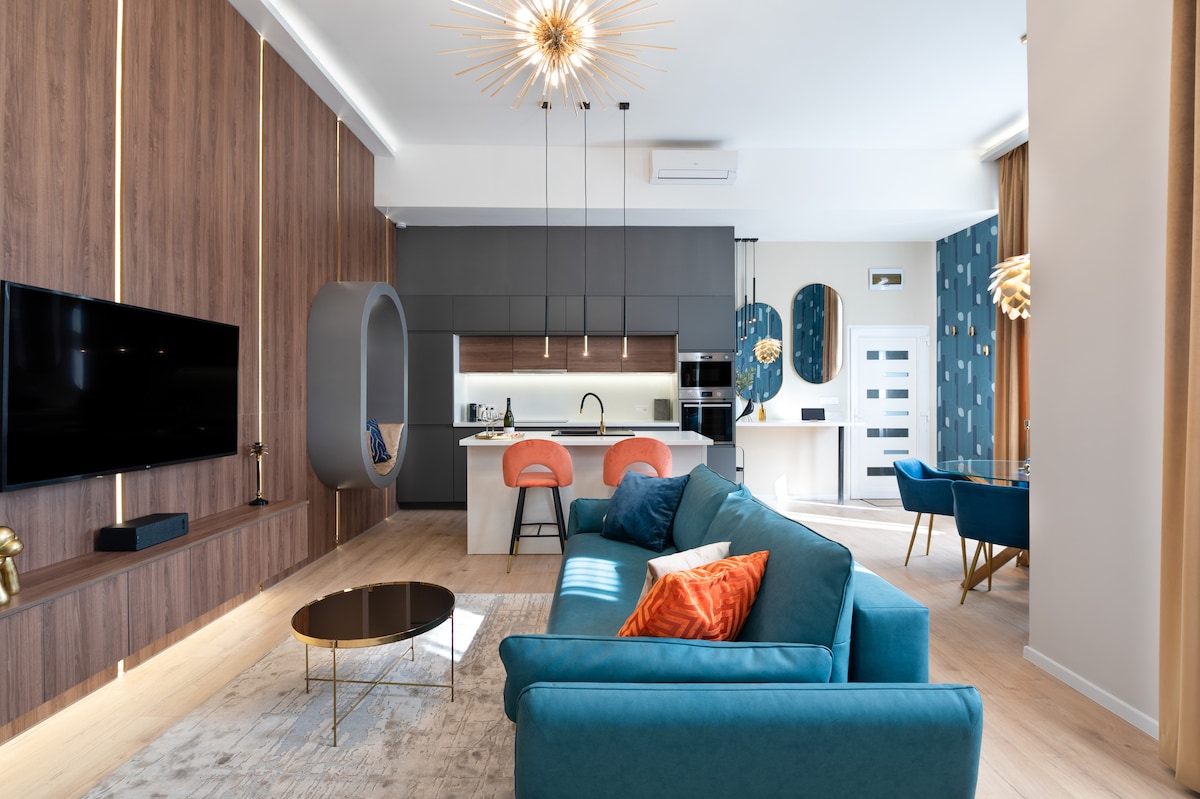 A spacious living area features a modern teal sofa with decorative pillows, contrasted by wooden paneling. A sleek kitchen island with bar stools is visible in the background, along with a dining table for four. A large flat-screen TV is mounted on the wall.