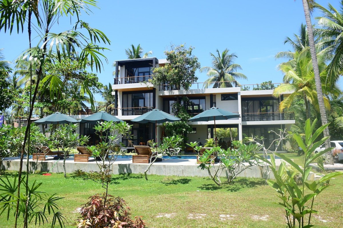 A modern, multi-level building is surrounded by lush greenery, featuring large glass windows that allow natural light to flow in. The outdoor area includes several lounge chairs shaded by umbrellas, providing a relaxed atmosphere near a tranquil pool.