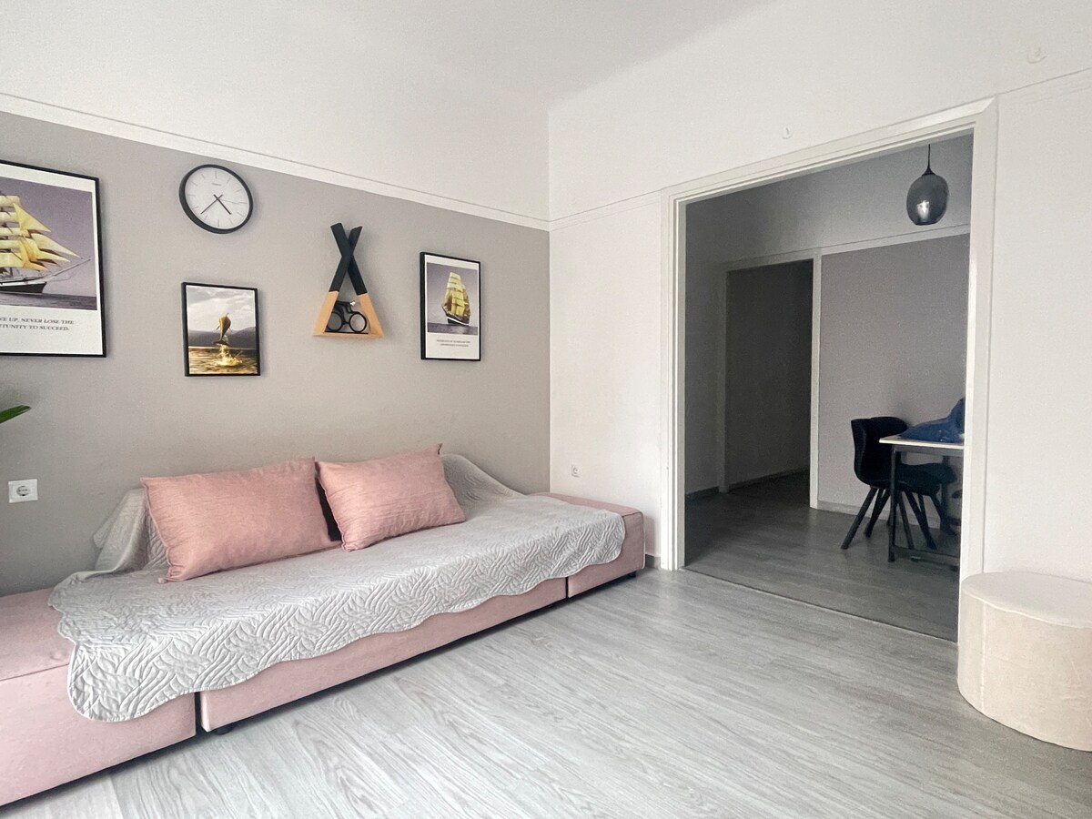 A light-filled living area features a pink sofa with a textured blanket draped over it. Artwork is displayed on the wall, including framed photographs and a clock. A hallway leads to a dining area, observed through an open doorway.