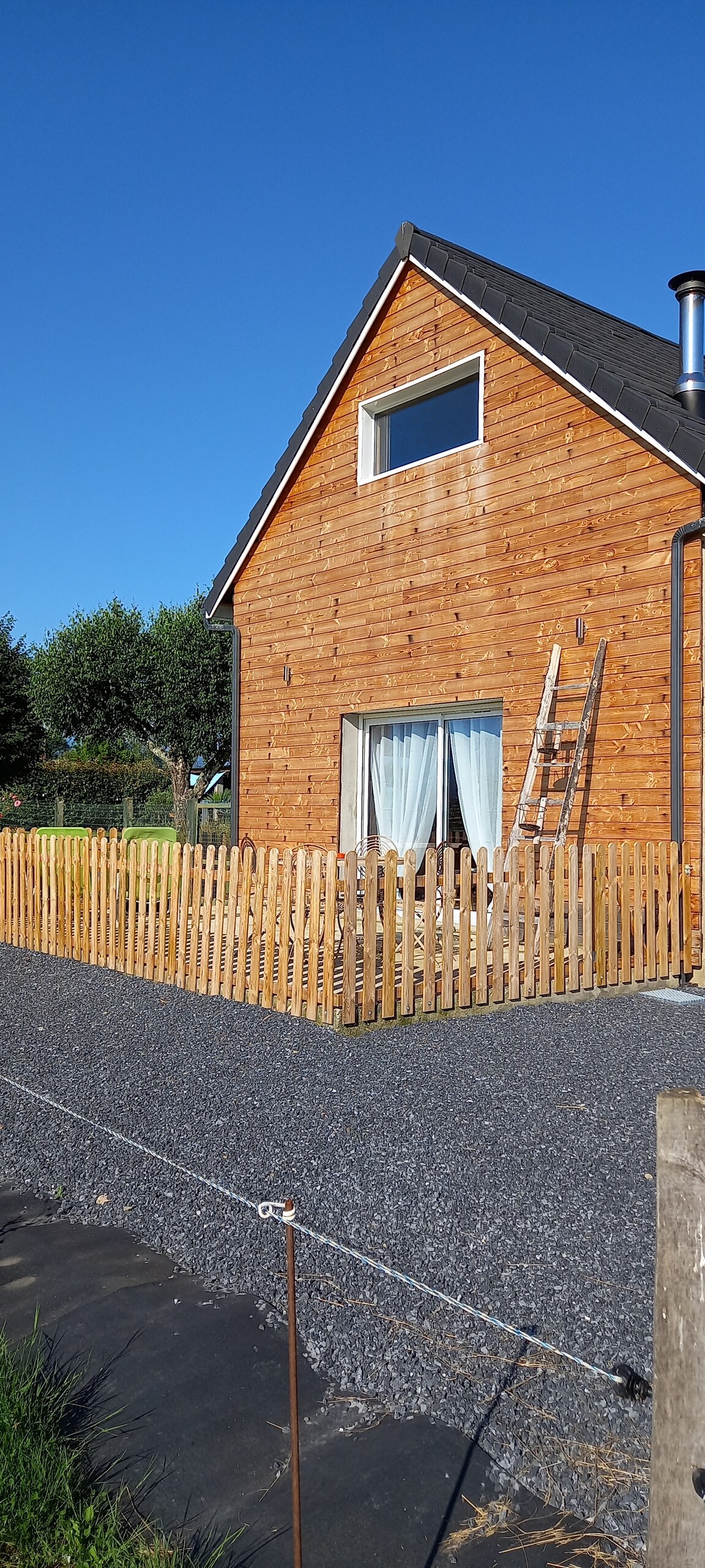 A charming renovated barn features a wooden exterior and a large window allowing natural light to enter. A wooden fence surrounds the property, offering a sense of privacy. A ladder rests against the side of the building, hinting at ongoing maintenance. Clear blue skies enhance the setting.