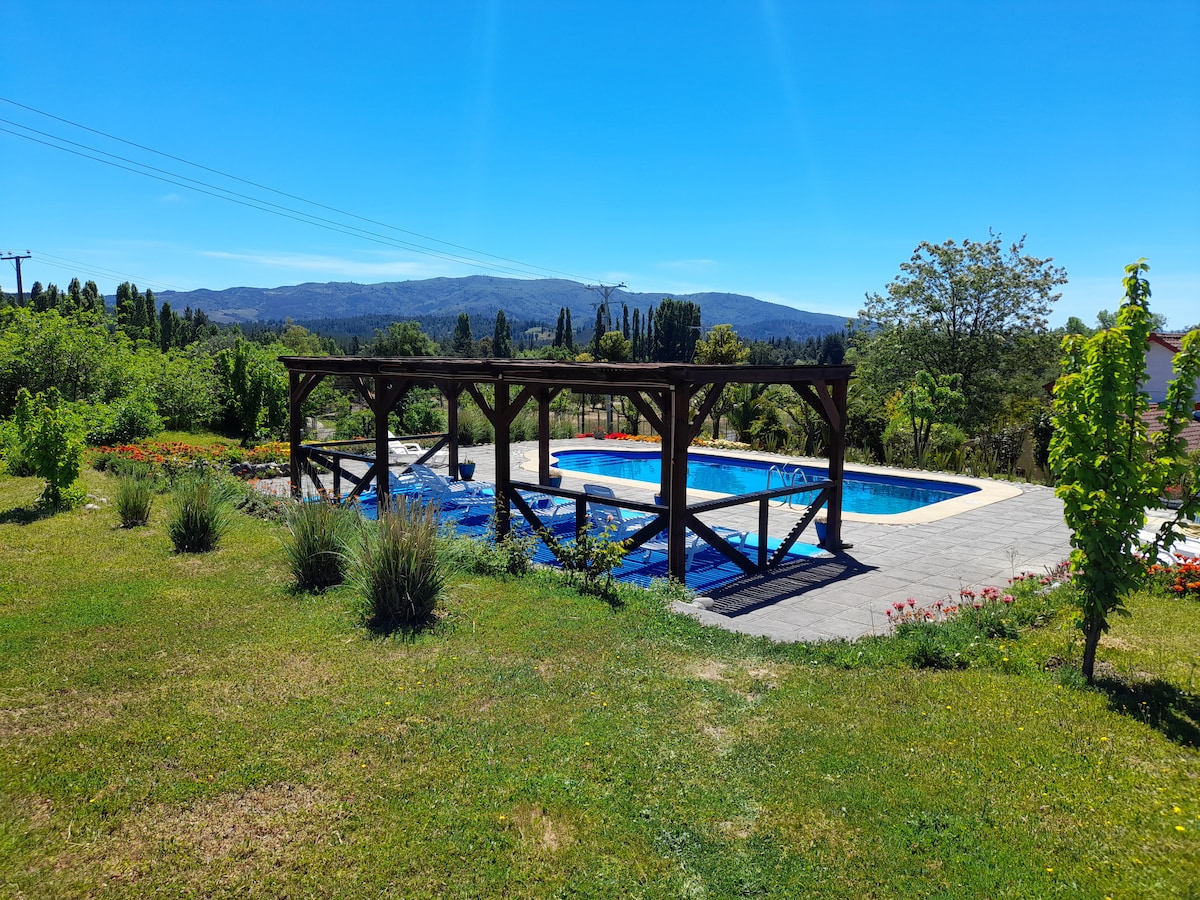 A serene outdoor space features two swimming pools surrounded by vibrant greenery and flowering plants. Wooden structures provide shade around the pools, and distant mountains create a peaceful backdrop under a clear blue sky.