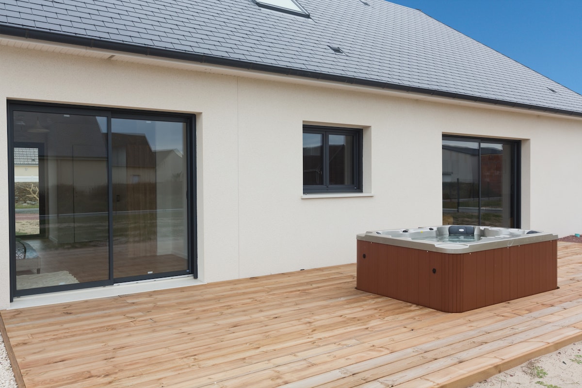 An outdoor wooden terrace features a spacious hot tub alongside large sliding glass doors that lead into the villa. The clean lines of the modern exterior are contrasted by a light-colored facade and a slate roof, highlighting the seamless connection to outdoor space.