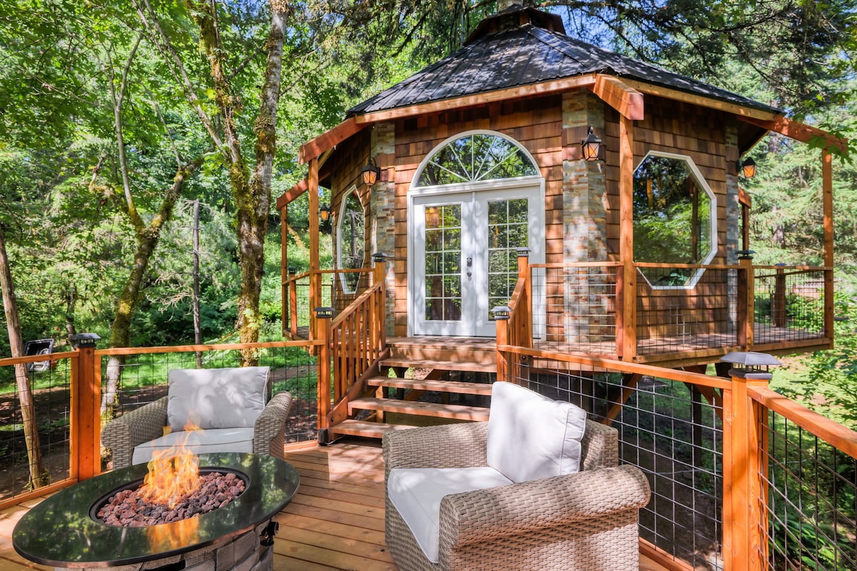 A charming treehouse features a unique octagonal design surrounded by lush greenery. A wooden deck extends from the structure, complemented by two comfortable chairs positioned around a circular fire pit, creating an inviting space for relaxation.