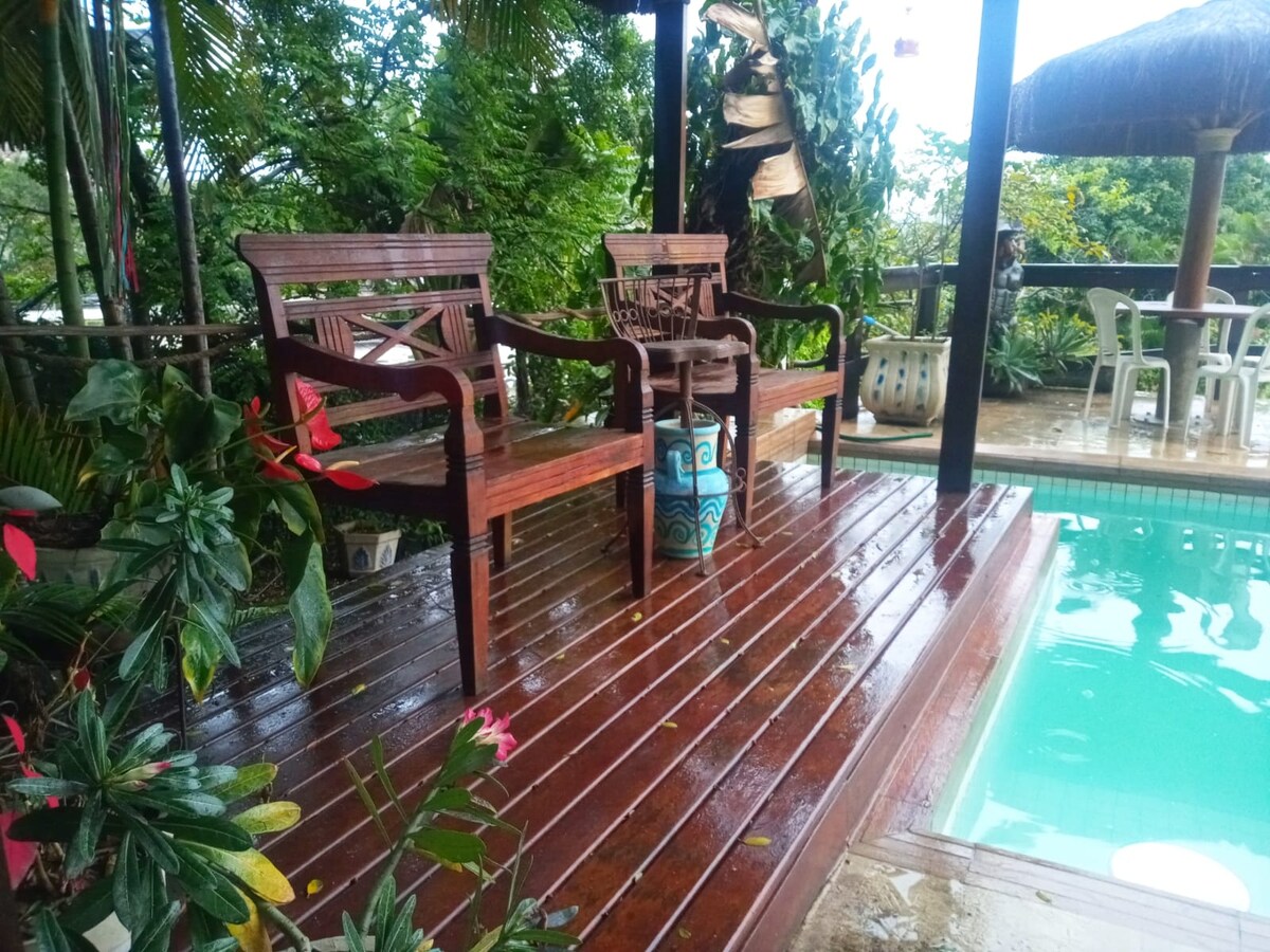 Two wooden chairs are positioned on a deck surrounded by lush greenery. A small table stands between them, and a pool can be seen nearby, reflecting the natural surroundings. The area is shaded by a thatched roof, providing a serene atmosphere.