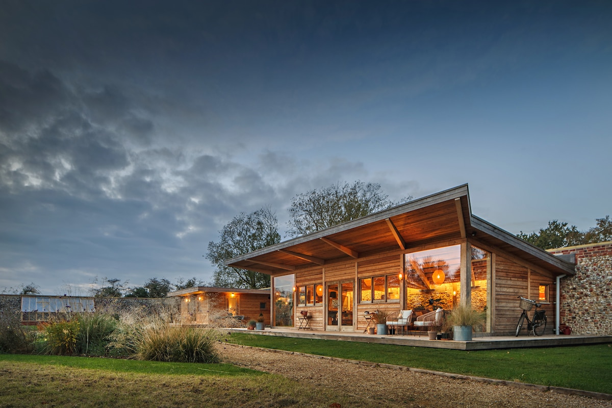 Two bespoke cabins are set amidst landscaped gardens, featuring large windows that reflect a warm glow. The exterior showcases a combination of wood and stone, framed by lush greenery and grass. A serene atmosphere is created by the evening light and soft, cloudy sky.