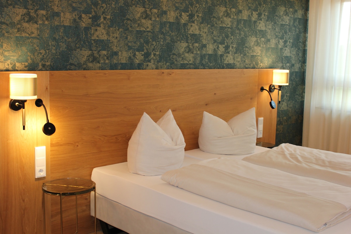 A bright and inviting room features a wooden headboard with two stylish wall lights. The spacious bed is made with crisp white linens and two decorative pillows. Soft natural light filters through sheer curtains, accentuating the textured blue wall behind the bed.