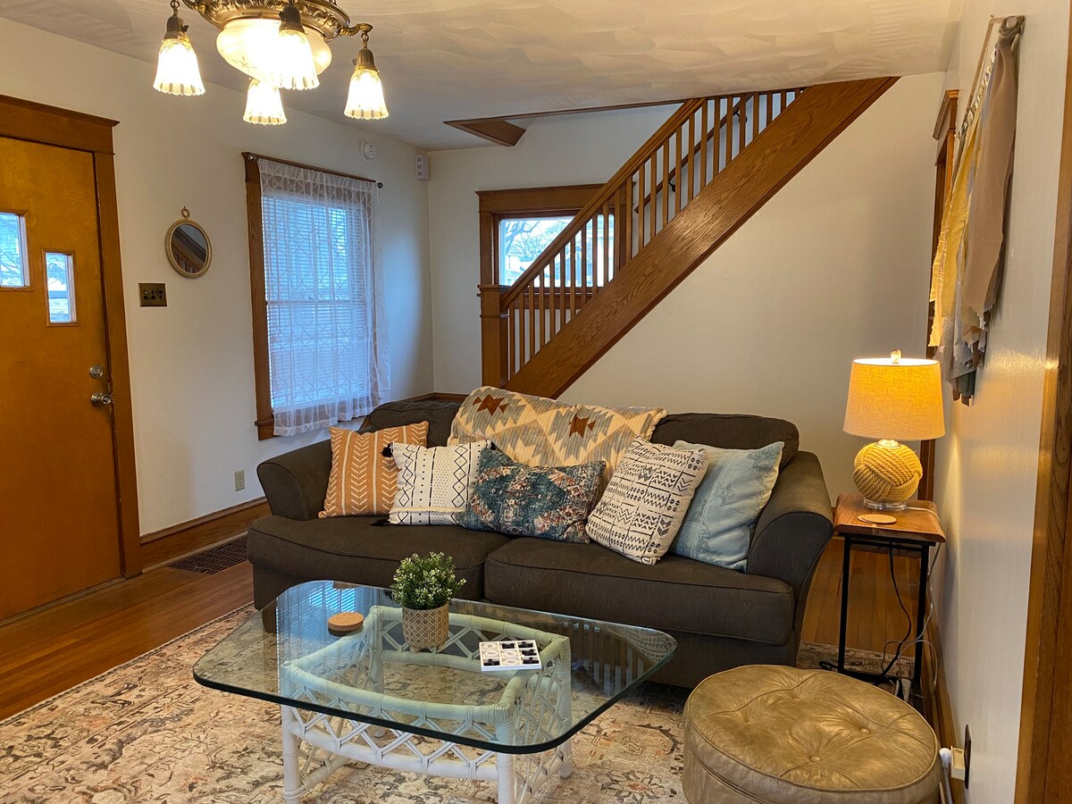 The living room features a comfortable sofa adorned with decorative pillows. A glass coffee table sits in front of the sofa, accompanied by a small lamp. Natural light enters through a nearby window, and a staircase leads to the upper floor, adding to the space's charm.