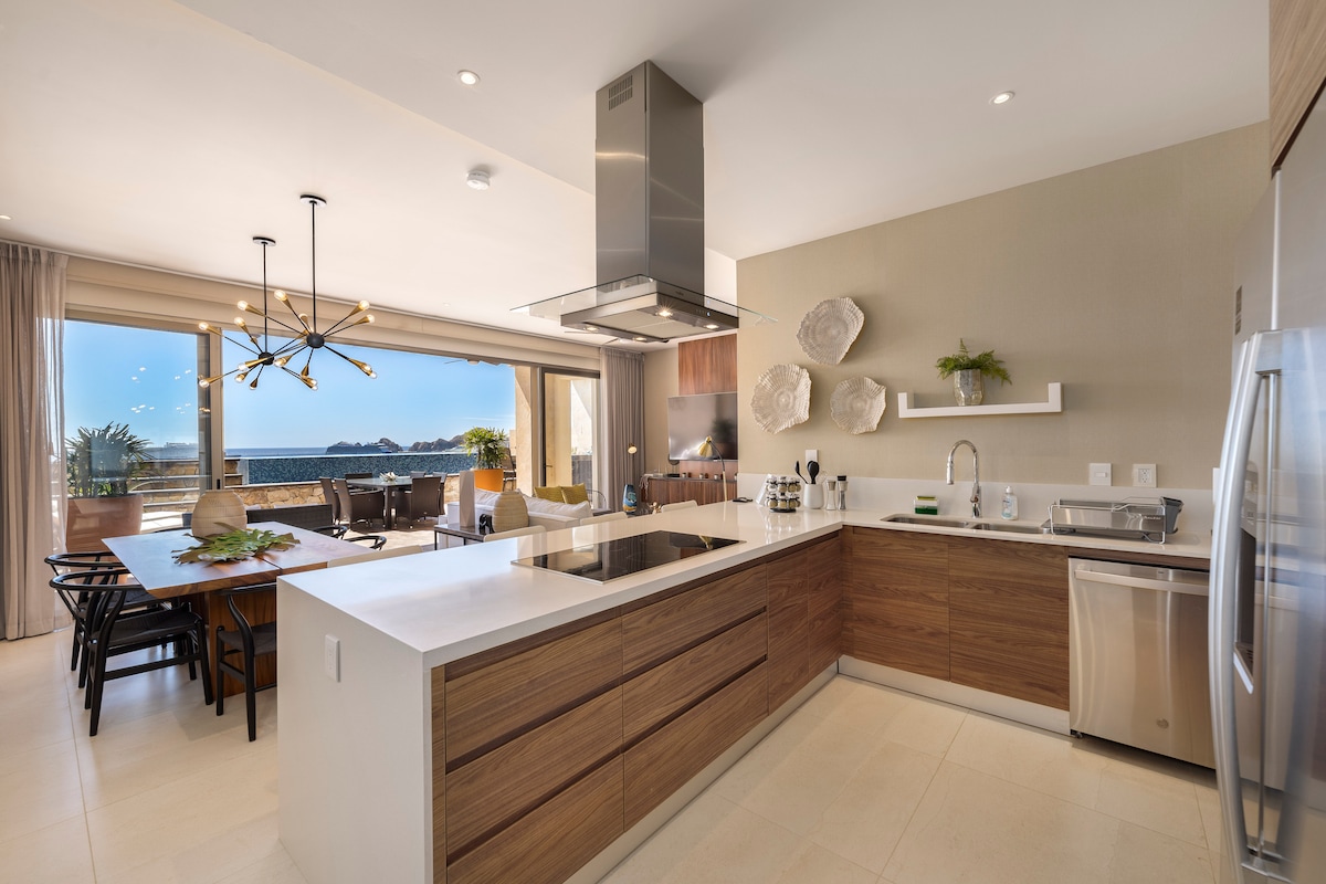 A modern kitchen is featured with sleek wooden cabinetry and stainless steel appliances. An island with a cooktop is positioned at the center. Expansive glass doors lead to a dining area and outdoor terrace, providing ample natural light and views of the surroundings.