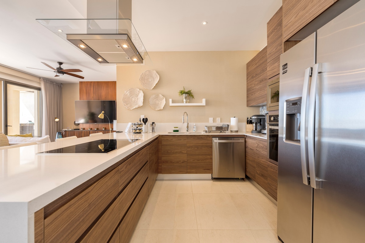 A modern kitchen is presented with sleek wooden cabinetry and stainless steel appliances. A large island with a smooth countertop provides ample workspace. Natural light fills the space through sliding glass doors, and a decorative piece is displayed on the wall above the sink.